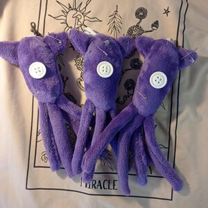May include: Three plush purple octopus toys with white button eyes and safety pin accents. Each toy has eight tentacles and small ear-like appendages. The toys are displayed against a light-colored background.