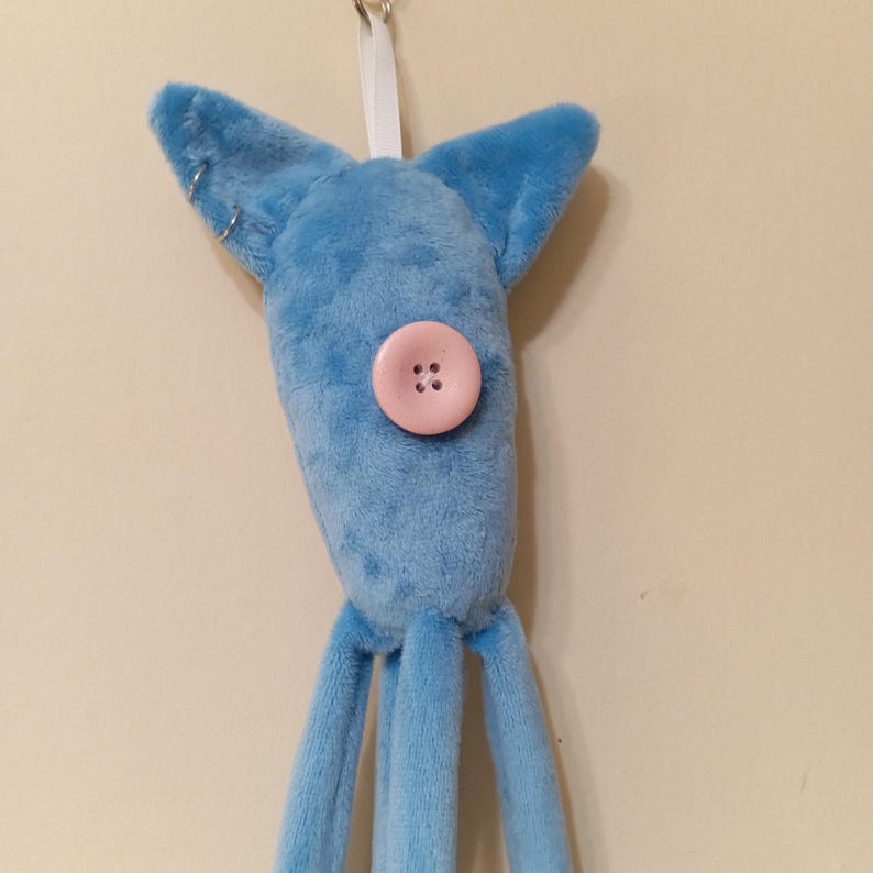 May include: A plush, light blue creature with large ears and three dangling appendages. A pink button with four holes serves as a facial feature. The toy is suspended by a white ribbon.