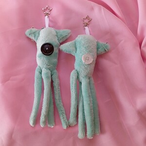 May include: Two plush, teal-colored octopus keychains with star-shaped clasps. Each keychain features a button on the body and three dangling tentacles. One has a dark button, the other a white button. The background is a pink fabric.