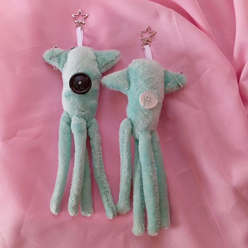May include: Two plush, teal-colored octopus keychains with star-shaped clasps. Each keychain features a button on the body and three dangling tentacles. One has a dark button, the other a white button. The background is a pink fabric.