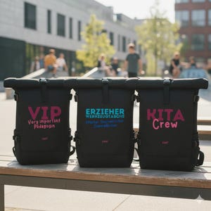 May include: Three black roll-top backpacks with text in pink, teal, and purple. The first backpack reads "VIP Very important Pädagogin". The second backpack reads "ERZIEHER Werkzeugtasche". The third backpack reads "KITA Crew". All three backpacks are on a wooden bench.