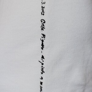 May include: White fabric with black embroidered text that reads "13.3.2012 Greta Pigatto - 4/pinta 9 mm"