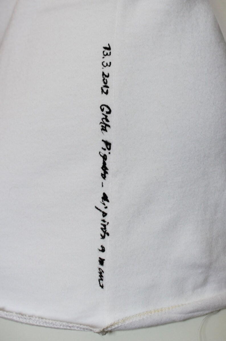 May include: White fabric with black embroidered text that reads "13.3.2012 Greta Pigatto - 4/pinta 9 mm"