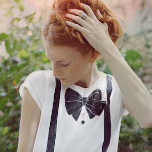 May include: A young woman with red hair is wearing a white t-shirt with a black bow tie and suspenders. The bow tie has a pink heart design in the center.