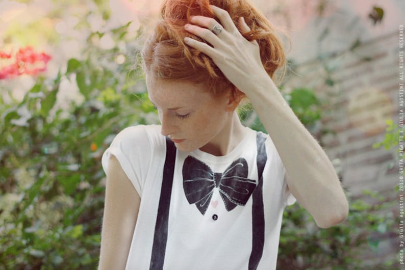 May include: A young woman with red hair is wearing a white t-shirt with a black bow tie and suspenders. The bow tie has a pink heart design in the center.
