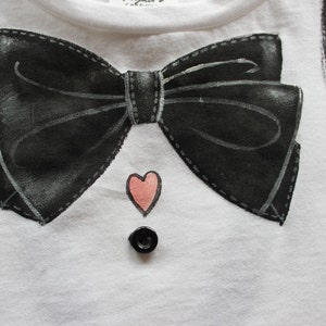 May include: White t-shirt with a black bow tie and a pink heart design. The shirt also has a black button.