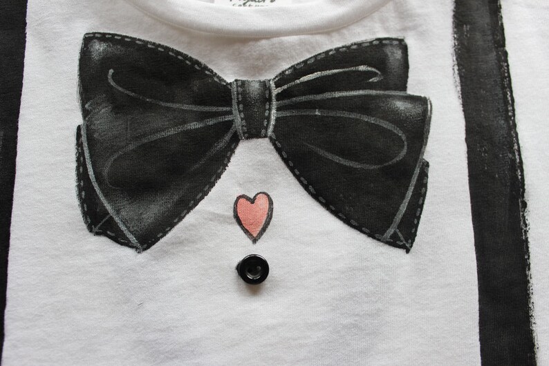May include: White t-shirt with a black bow tie and a pink heart design. The shirt also has a black button.