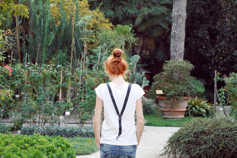 May include: A woman with red hair wearing a white t-shirt with black suspenders and blue jeans stands in a garden setting.