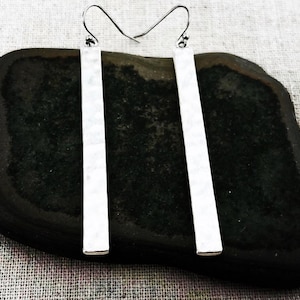 May include: A pair of silver bar earrings with a hammered texture. The earrings are rectangular and hang from a simple hook. The earrings are displayed on a dark, textured surface.