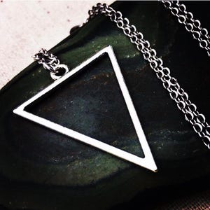 May include: Silver-toned necklace featuring a geometric triangle pendant. The pendant is a hollow, open triangle shape. The chain is a delicate, linked design. The necklace is displayed on a dark, textured surface, creating a contrast with the bright metal.