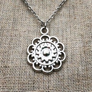 Puede incluir: Collar con colgante plateado con dise&ntilde;o floral. El colgante tiene un centro circular con un dise&ntilde;o de estrella, rodeado por un borde festoneado. La cadena es de estilo eslab&oacute;n delicado, y el fondo es una tela texturizada y neutra.