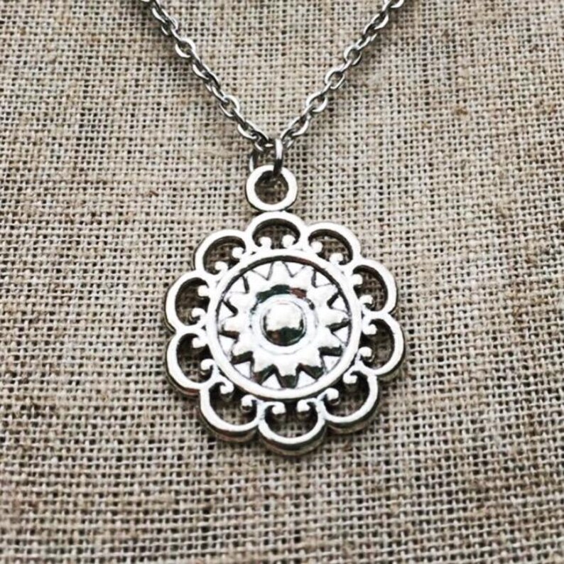 Puede incluir: Collar con colgante plateado con dise&ntilde;o floral. El colgante tiene un centro circular con un dise&ntilde;o de estrella, rodeado por un borde festoneado. La cadena es de estilo eslab&oacute;n delicado, y el fondo es una tela texturizada y neutra.