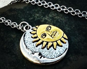May include: A silver-toned necklace featuring a pendant with a gold sun face over a silver moon. The sun has a smiling face with rays extending outward. The moon has a textured surface and a small face. The chain is a delicate link style.