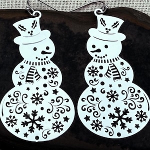 May include: A pair of silver-toned metal earrings shaped like snowmen. Each snowman wears a top hat with holly leaves and berries, a striped scarf, and features intricate cut-out patterns of snowflakes, stars, and swirling filigree designs.
