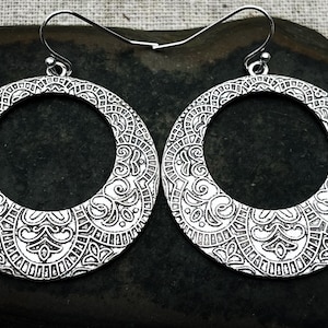 May include: A pair of silver hoop earrings with an intricate, ornate design. The earrings are circular and have a large, open center.