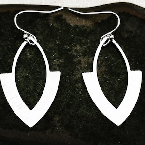 May include: A pair of silver earrings. Each earring has an open, elongated teardrop shape with a flat base. The earrings are suspended from a simple hook. The earrings are displayed against a dark background.