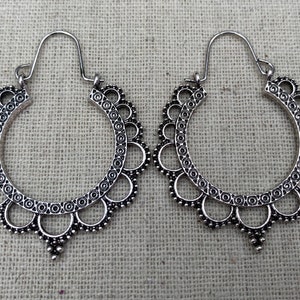 May include: A pair of silver-colored hoop earrings. Each earring features a scalloped edge with circular and dot patterns. The earrings have a hook closure and a decorative pendant at the bottom.