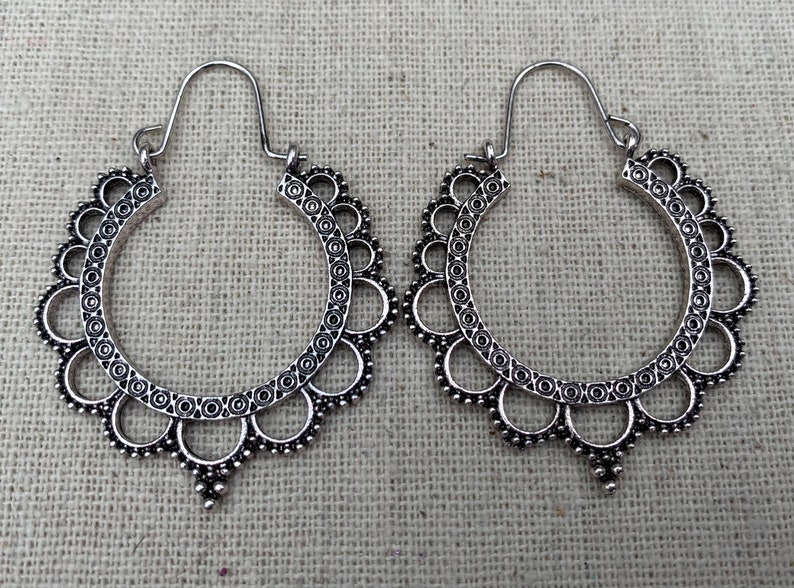 May include: A pair of silver-colored hoop earrings. Each earring features a scalloped edge with circular and dot patterns. The earrings have a hook closure and a decorative pendant at the bottom.