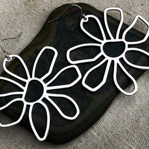 May include: A pair of silver-colored flower-shaped earrings. Each earring features a stylized floral design with open petals and a dark center. The earrings are displayed on a dark, textured surface.