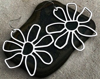 May include: A pair of silver-colored flower-shaped earrings. Each earring features a stylized floral design with open petals and a dark center. The earrings are displayed on a dark, textured surface.