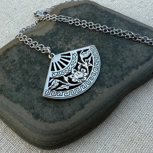 May include: A silver-colored pendant necklace featuring a fan-shaped design with floral and geometric patterns. The pendant is suspended from a delicate chain. The necklace is displayed on a dark gray stone.