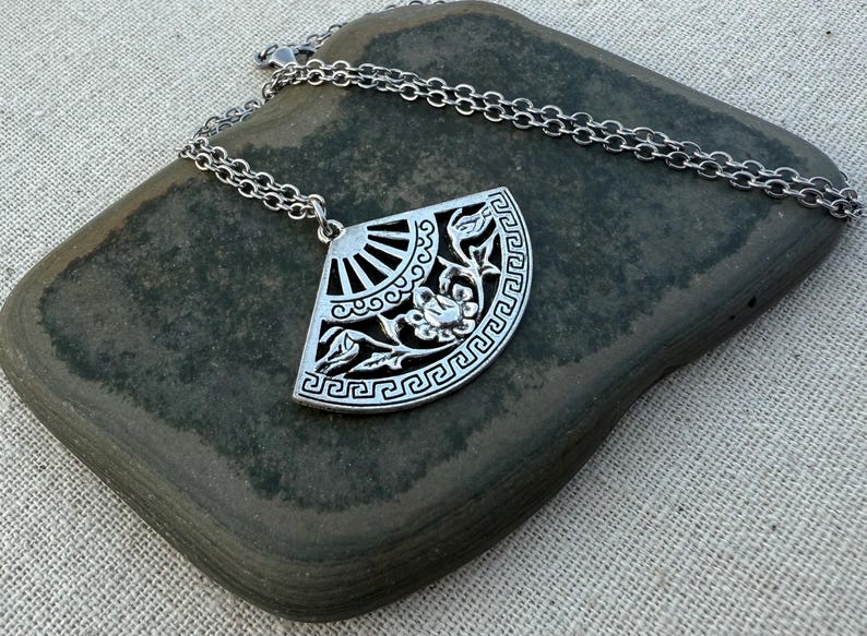 May include: A silver-colored pendant necklace featuring a fan-shaped design with floral and geometric patterns. The pendant is suspended from a delicate chain. The necklace is displayed on a dark gray stone.