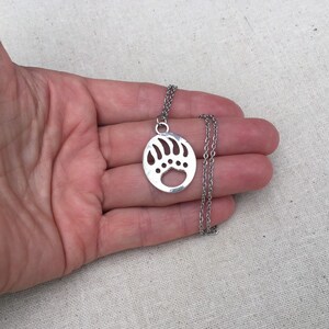May include: A silver necklace with a bear paw print pendant. The pendant is a circle with a cut-out bear paw print.
