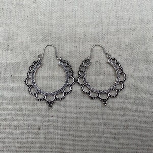 May include: A pair of silver-toned hoop earrings. Each earring features a scalloped edge design with small circular details and a dangling teardrop shape at the bottom. The earrings are displayed on a neutral-colored fabric background.