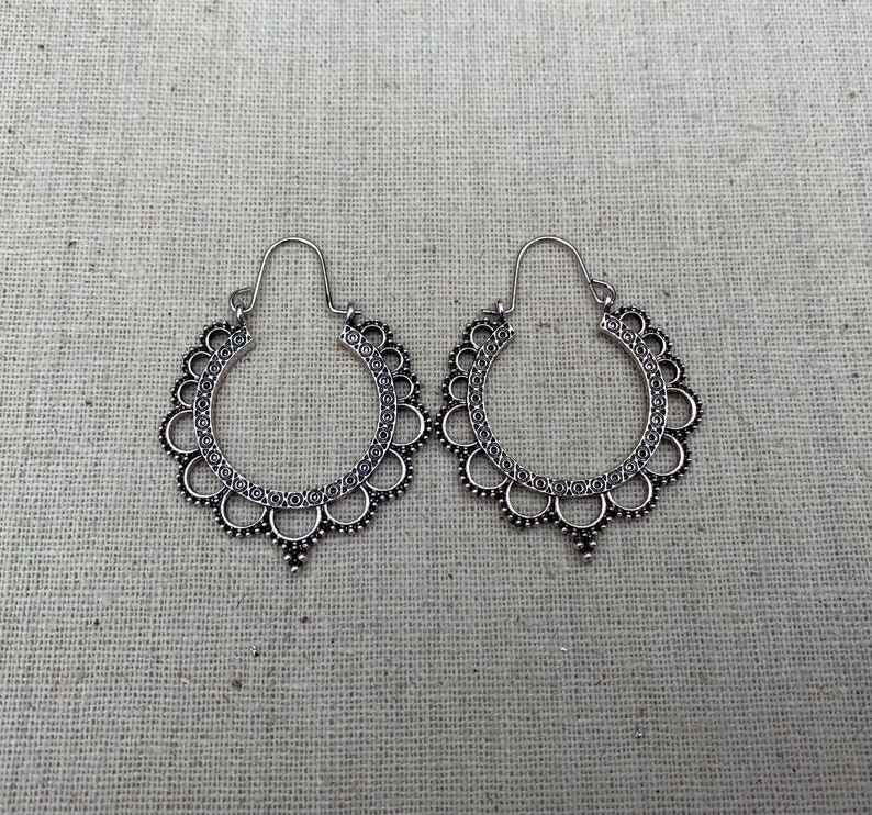 May include: A pair of silver-toned hoop earrings. Each earring features a scalloped edge design with small circular details and a dangling teardrop shape at the bottom. The earrings are displayed on a neutral-colored fabric background.