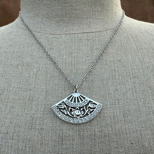 May include: Silver-toned pendant necklace featuring a fan-shaped charm. The charm has intricate cut-out designs, including floral and geometric patterns. The necklace hangs on a delicate chain, creating an elegant accessory.