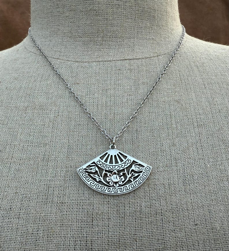 May include: Silver-toned pendant necklace featuring a fan-shaped charm. The charm has intricate cut-out designs, including floral and geometric patterns. The necklace hangs on a delicate chain, creating an elegant accessory.