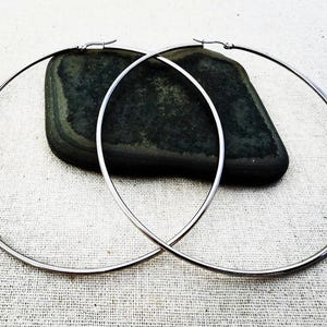 May include: Two large, silver-colored hoop earrings. The hoops are round and have a simple, minimalist design. The earrings are displayed on a light-colored, textured surface with a dark, oblong stone in the background.