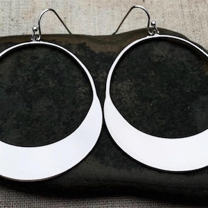 May include: A pair of silver-colored hoop earrings. The earrings have a unique design, with a flat, curved bottom and a rounded top. They are displayed on a dark, textured surface.
