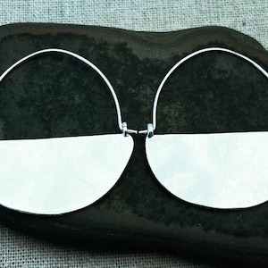 May include: A pair of silver-toned half-moon earrings. Each earring features a curved wire that connects to a flat, semi-circular shape. The earrings are displayed on a dark, textured surface, showcasing their minimalist design and metallic sheen.