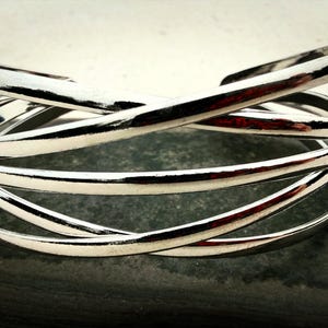 May include: A polished silver-tone metal cuff bracelet featuring multiple overlapping, thin, rounded bands that create a layered, crisscross design. The bracelet has a smooth, reflective surface and an open-ended structure for easy wear.