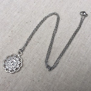 Puede incluir: Un collar plateado con un colgante floral. El colgante tiene un dise&ntilde;o calado detallado con un elemento circular central. La cadena es delicada y tiene un cierre de mosquet&oacute;n. El collar se muestra sobre una tela de color neutro.