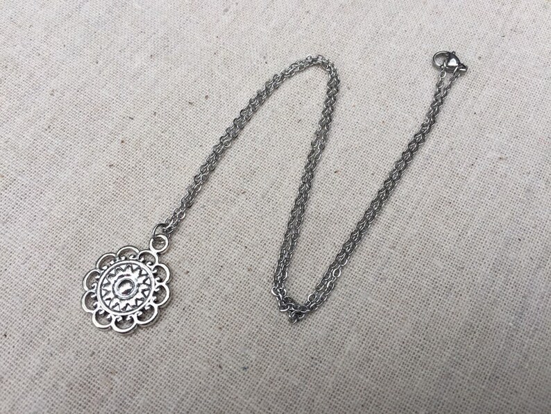 Puede incluir: Un collar plateado con un colgante floral. El colgante tiene un dise&ntilde;o calado detallado con un elemento circular central. La cadena es delicada y tiene un cierre de mosquet&oacute;n. El collar se muestra sobre una tela de color neutro.