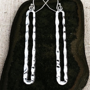 May include: Silver-toned, hammered-metal earrings. The earrings are long, rectangular, and have a textured surface with a cutout design. They hang from a simple hook. The earrings are displayed on a dark, textured surface.