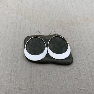 Puede incluir: Un par de pendientes de aro plateados con una base plana en forma de media luna. Los pendientes se muestran sobre una piedra gris oscura sobre un fondo de tela neutra. Dise&ntilde;o sencillo y moderno.