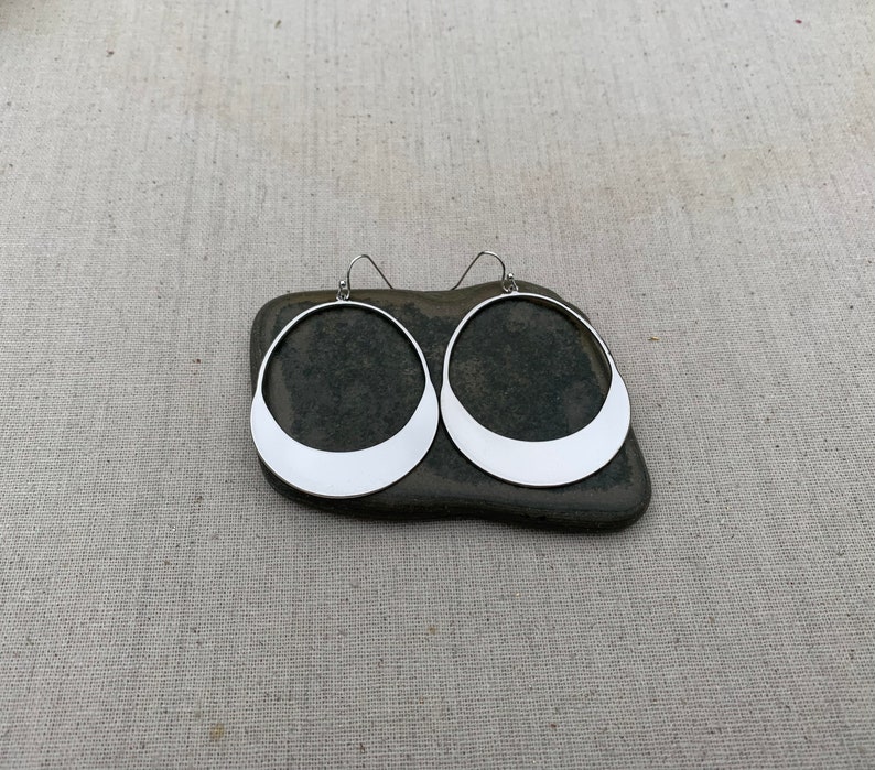 Puede incluir: Un par de pendientes de aro plateados con una base plana en forma de media luna. Los pendientes se muestran sobre una piedra gris oscura sobre un fondo de tela neutra. Dise&ntilde;o sencillo y moderno.