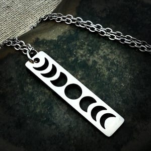May include: Silver-toned necklace featuring a rectangular pendant with cut-out moon phase designs. The pendant is connected to a delicate chain. The necklace is displayed on a dark, textured surface, creating a contrast with the bright metal.