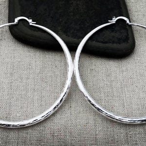 May include: A pair of silver hoop earrings with a hammered texture. The earrings are round and have a simple clasp closure. They are displayed on a neutral linen surface with a dark, textured object in the background.