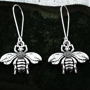 May include: A pair of silver bee earrings. Each earring has a detailed bee pendant with wings and a flower-like centre, suspended from a long, curved ear wire. The earrings are shown against a dark background.