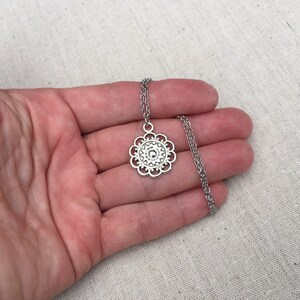 Puede incluir: Collar plateado con un colgante floral calado. El colgante est&aacute; suspendido de una fina cadena. El collar se muestra sobre un fondo neutro.