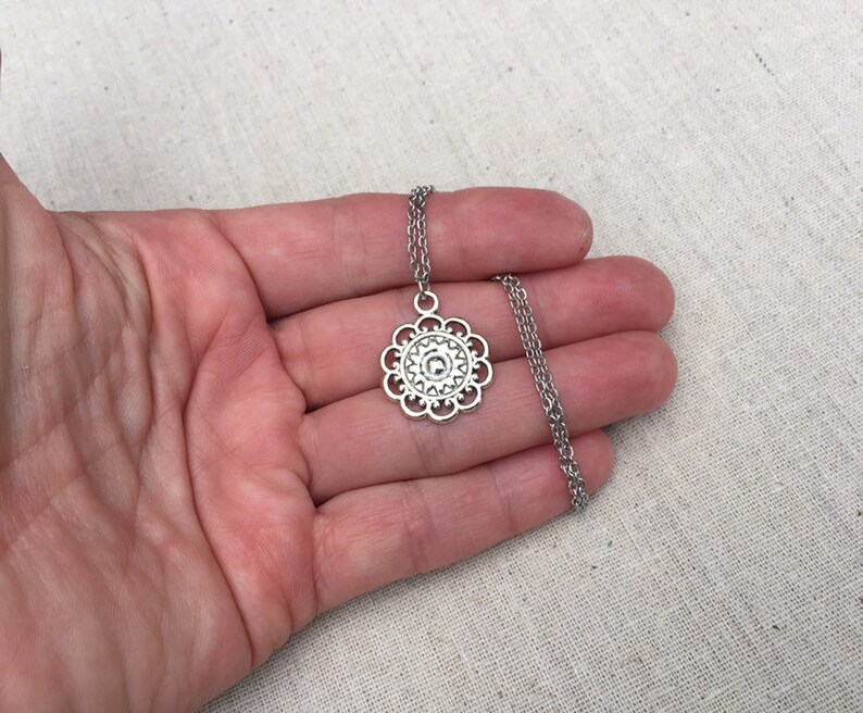 Puede incluir: Collar plateado con un colgante floral calado. El colgante est&aacute; suspendido de una fina cadena. El collar se muestra sobre un fondo neutro.