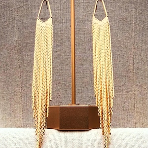 Peut inclure: Une paire de longues boucles d'oreilles pendantes dorées dotées d'une monture supérieure géométrique et angulaire, reliée à une frange dense et tombante de fines chaînes métalliques qui pendent verticalement, créant un aspect texturé.