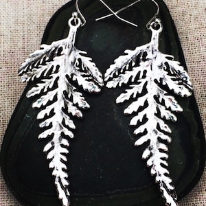 May include: A pair of silver-colored leaf-shaped earrings. The earrings are detailed with intricate veining and have a hook closure. The earrings are displayed on a dark, textured surface.