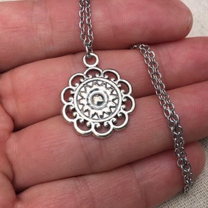 Puede incluir: Collar plateado con colgante floral. El colgante tiene un centro circular con un dise&ntilde;o de rayos de sol y un borde festoneado. La cadena es de estilo delicado y enlazado.