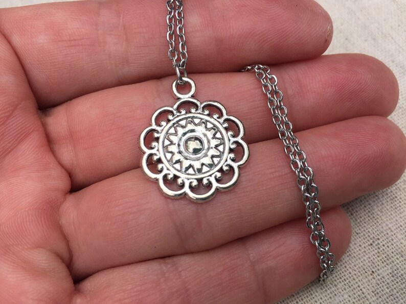 Puede incluir: Collar plateado con colgante floral. El colgante tiene un centro circular con un dise&ntilde;o de rayos de sol y un borde festoneado. La cadena es de estilo delicado y enlazado.