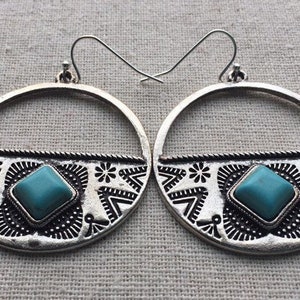 May include: A pair of silver-toned hoop earrings with a turquoise square stone at the center. The earrings have a decorative pattern along the bottom edge, featuring floral and geometric designs. The earrings are displayed on a neutral fabric background.